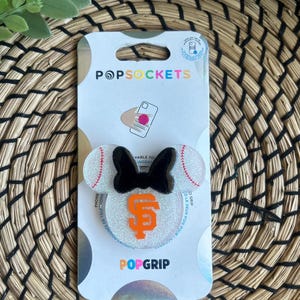 May include: A white and silver glitter phone grip with a black bow and a baseball design. The grip has the San Francisco Giants logo in orange. The text "PopGrip" is printed on the packaging.