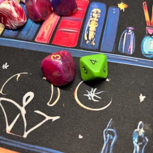 Puede incluir: Una colección de dados de juego de varias caras en diferentes colores, incluyendo magenta, morado y verde lima. Los dados presentan números en dorado y negro. Algunos dados tienen un acabado jaspeado o brillante, mientras que otros son de color sólido.