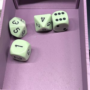 May include: A light green set of polyhedral dice resting inside a lavender-colored rectangular tray. The dice include a six-sided die with black pips, a four-sided die with black numbers, and two other multi-sided dice with black numerical markings.