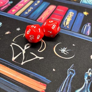 May include: Two bright red ten-sided dice with white numbers resting on a black surface featuring a printed illustration of colorful books and various mystical symbols including a crescent moon and abstract line art designs.