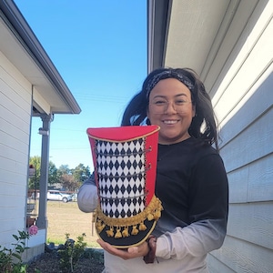 May include: A person is holding a red, black, and gold top hat with a black and white diamond pattern. The hat has gold tassels and a black brim.
