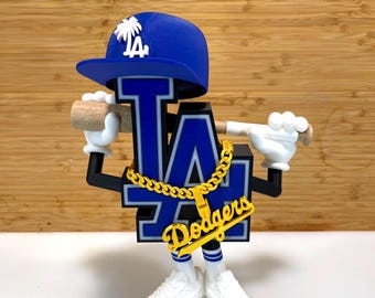 Könnte beinhalten: Eine stilisierte Dodgers-Logo-Figur mit einer blauen Baseballkappe mit einer weißen Palme. Die Figur trägt eine goldene Kette mit einem "Dodgers"-Anhänger, weiße Turnschuhe und hält einen Baseballschläger. Der Hintergrund ist hellbraunes Holz.