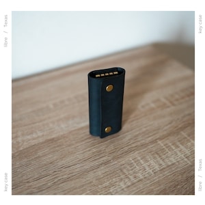 May include: A black leather key case with gold accents and a snap closure. The case is sitting on a wooden surface.
