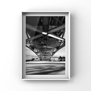 May include: Black and white photograph of a person cycling beneath a bridge. The bridge is constructed from metal and has a complex design. The photo is taken from below the bridge, looking upwards.