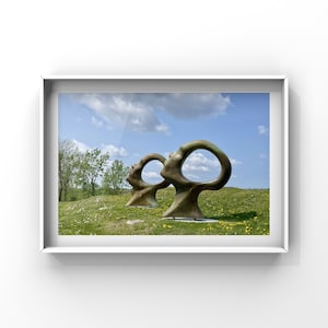 May include: Two abstract bronze sculptures in a grassy field with a blue sky and white clouds in the background. The sculptures are shaped like large, open circles.