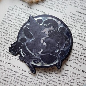 Puede incluir: Pegatina en blanco y negro que representa a una mujer montada a caballo, con un diseño de media luna. La pegatina está colocada en una página de un libro antiguo con texto y un collar de perlas.