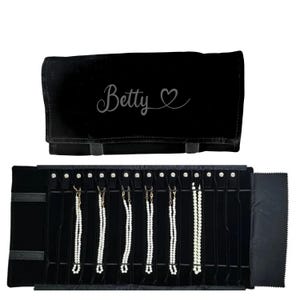 May include: Black velvet jewelry roll with the name "Betty" and a heart design. The roll is open, displaying several necklaces with white beads. The roll has a flap to close and secure the jewelry.