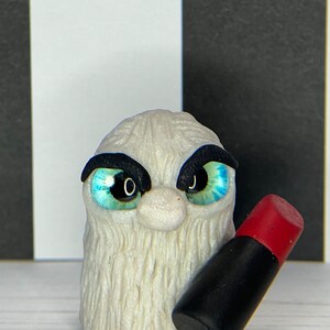 Puede incluir: Una criatura blanca y peluda con cejas negras y grandes ojos azules sostiene un tubo de lápiz labial negro y rojo.