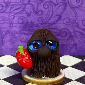 May include: A whimsical, handmade sculpture of a creature with dark brown fur, large blue eyes, and a dark brown nose. It holds a bright red strawberry. The sculpture sits on a small, round base, against a purple floral background.