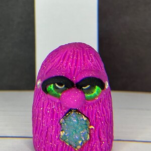 Peut inclure: Une petite créature poilue violette avec des yeux verts et un gros bijou bleu scintillant dans sa bouche. La créature a une expression renfrognée.