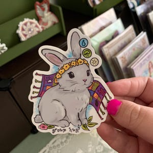 May include: A sticker featuring a gray bunny with a floral crown and a colorful patterned scarf. The sticker has the text "group hug?" and symbols representing water, earth, and air. The bunny is sitting on a bed of flowers.