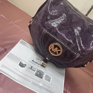 May include: A purple Michael Kors handbag with a snakeskin pattern. The bag features a gold-tone MK logo and hardware. A certificate of authenticity is visible, with the words "Authentic Handbags - Michael Kors".