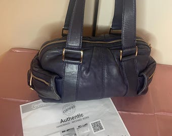 May include: A navy blue leather handbag with gold-toned hardware, featuring two exterior zippered pockets and dual top handles. The bag is accompanied by a white "Authentic" certification document. The bag's design includes a top zipper closure.