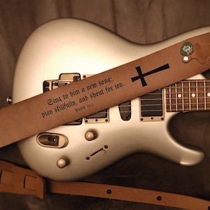 May include: A brown leather guitar strap with a black cross and the text "Sing to him a new song; play skillfully, and shout for joy. Psalm 33:3" printed on it. The strap is on a silver electric guitar.