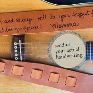May include: A brown leather guitar strap with a handwritten message that reads "I am and always will be your biggest fan! I love you forever! Momma". The strap is on a wooden acoustic guitar with a soundhole that has a beige paper circle with the text "send us your actual handwriting".