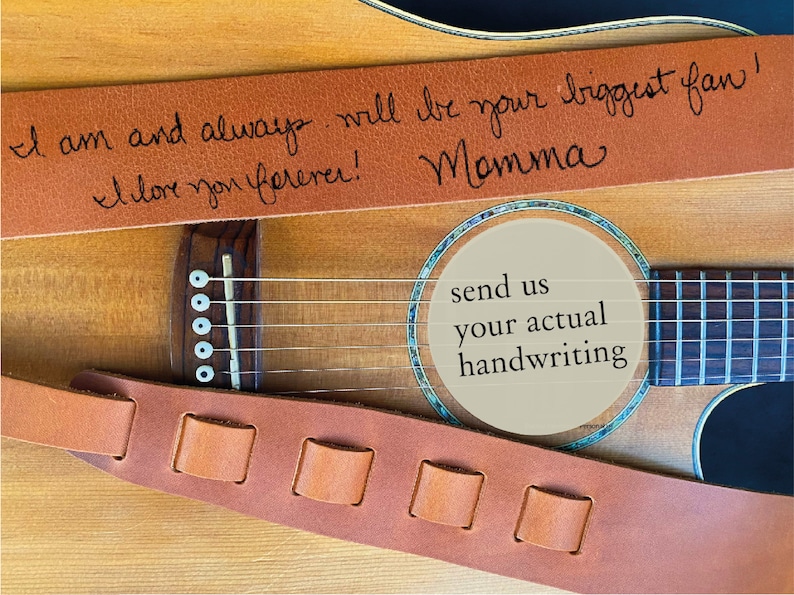 May include: A brown leather guitar strap with a handwritten message that reads "I am and always will be your biggest fan! I love you forever! Momma". The strap is on a wooden acoustic guitar with a soundhole that has a beige paper circle with the text "send us your actual handwriting".