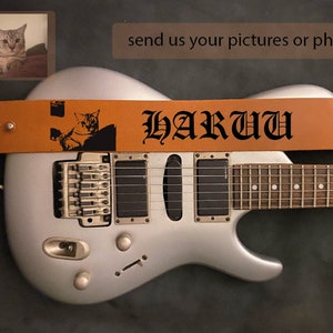 May include: A silver electric guitar with a brown leather guitar strap. The strap has a black cat silhouette and the text "HARU" in black gothic lettering.