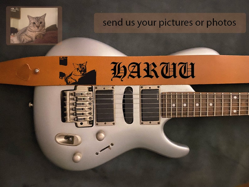May include: A silver electric guitar with a brown leather guitar strap. The strap has a black cat silhouette and the text "HARU" in black gothic lettering.