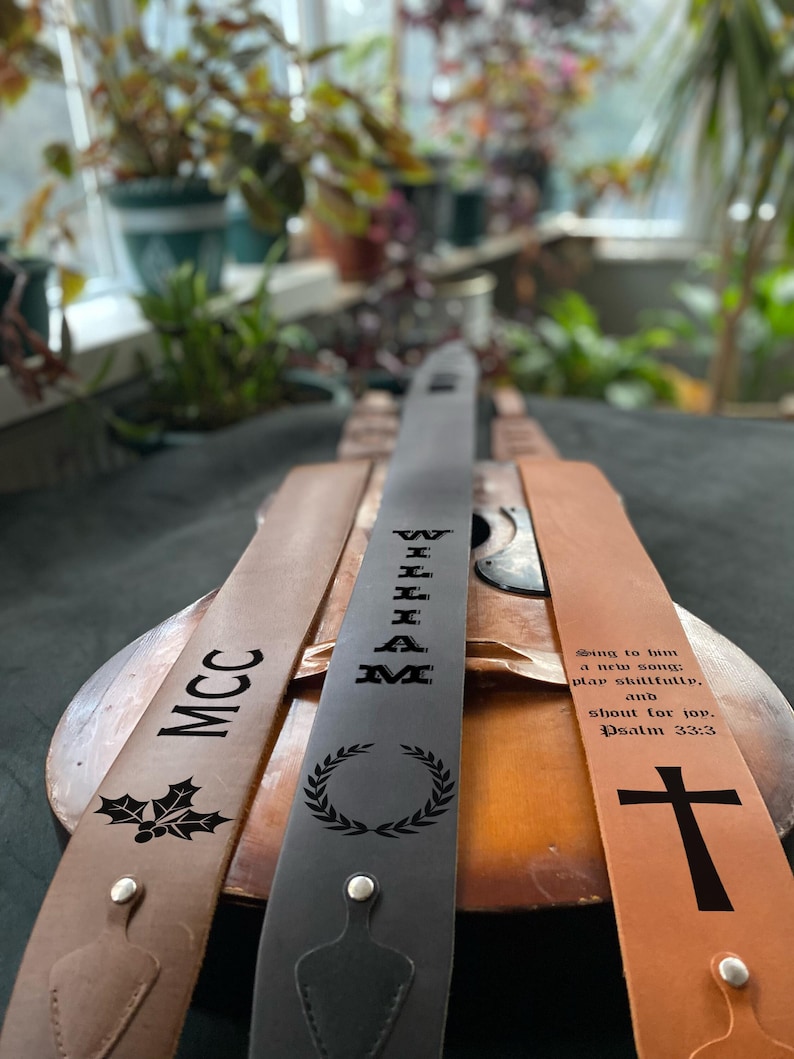 May include: Three leather guitar straps with different designs. The first strap is brown with a holly design and the text "MCC". The second strap is black with a laurel wreath design and the text "WILLIAM". The third strap is brown with a cross design and the text "Sing to him a new song: play skillfully, and shout for joy. Psalm 33:3".