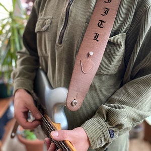May include: A brown leather guitar strap with the name "FJTL" embossed in black lettering. The strap is worn by a person playing a silver electric guitar.