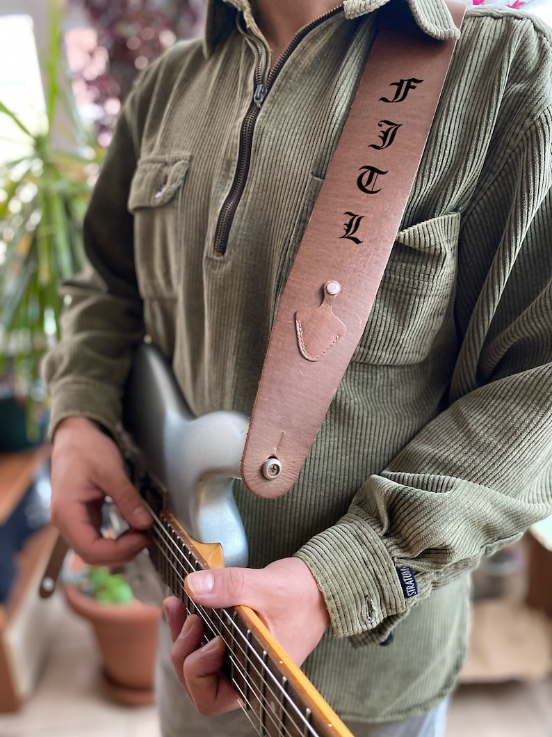 May include: A brown leather guitar strap with the name "FJTL" embossed in black lettering. The strap is worn by a person playing a silver electric guitar.