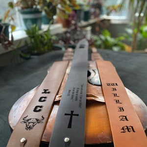 May include: Three guitar straps with different designs. One strap is brown with a bull's head and the text "HOGG". Another strap is black with a cross and the text "Jesus said to him, 'If you can believe, all things are possible.' Mark 9:23". The third strap is brown with the text "WILLIAM" in old English lettering.