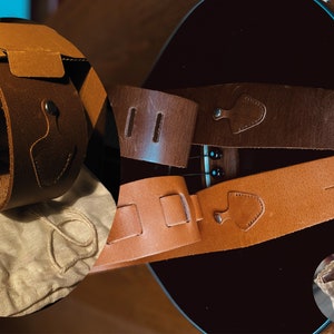May include: Two leather guitar straps, one brown and one tan, with a brown leather guitar strap in its packaging. The straps have adjustable buckles and are made of high-quality leather.