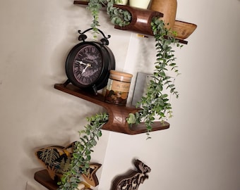 Puede incluir: Un conjunto de tres estantes de esquina de madera oscura montados en una pared. Los estantes sostienen varios artículos, incluyendo tres pequeñas plantas en macetas, un jarrón marrón alto, un reloj analógico negro, una vela en vaso y figuras de madera.