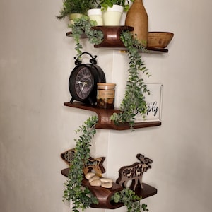 Peut inclure: Étagère d'angle en bois à trois niveaux, ornée d'objets décoratifs. L'étagère supérieure contient des plantes en pot, une bouteille marron et un bol en bois. L'étagère du milieu présente une horloge noire et un pot. L'étagère inférieure expose des découpes en bois et des pierres.