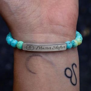 May include: A beaded bracelet featuring round, turquoise-coloured stones with natural veining and a central, rectangular silver-toned metal bar. The bar is engraved with a circular symbol and the text "Master Shifter" in a cursive script font.