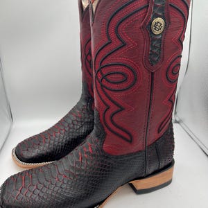 May include: A pair of cowboy boots with a striking design. The boots feature a combination of burgundy and black leather, with intricate red stitching and a snakeskin-like texture on the lower portion. A gold emblem is visible on the upper shaft.