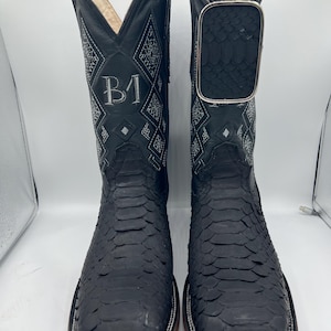 May include: A pair of black cowboy boots with a square toe and a textured snakeskin pattern. The boots have a decorative stitched design in white and the initials "B1" on the shaft. The boots have a silver buckle and a leather sole.