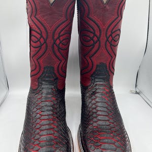 May include: A pair of cowboy boots with a square toe. The boots feature a burgundy leather upper with intricate black stitching and a black, textured lower section. The soles are light brown with a white and silver trim.