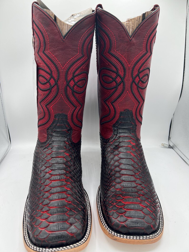 May include: A pair of cowboy boots with a square toe. The boots feature a burgundy leather upper with intricate black stitching and a black, textured lower section. The soles are light brown with a white and silver trim.