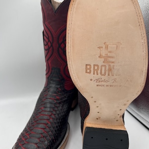 May include: A pair of cowboy boots with a snakeskin pattern in black and red. The boots have a square toe and a decorative stitch pattern on the shaft. The sole of the boot is visible, with the text "BRONZ" and "Exotic Boots Made in Mexico".