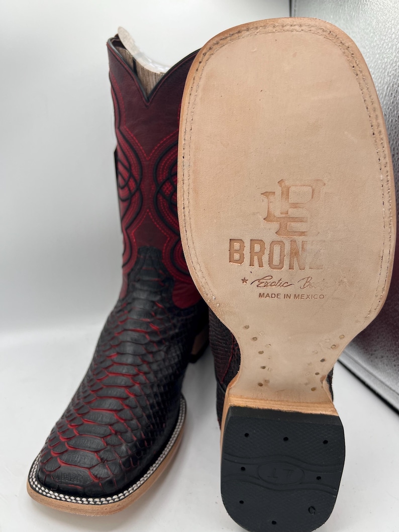 May include: A pair of cowboy boots with a snakeskin pattern in black and red. The boots have a square toe and a decorative stitch pattern on the shaft. The sole of the boot is visible, with the text "BRONZ" and "Exotic Boots Made in Mexico".