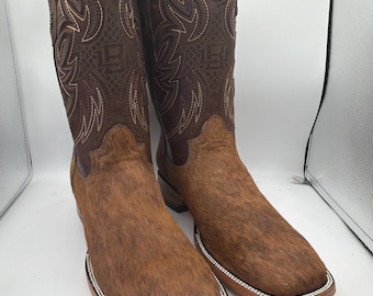 May include: A pair of brown leather cowboy boots with a square toe. The boots have a textured, fur-like appearance on the lower part and decorative stitching on the upper shaft. The boots have a white beaded trim around the sole.