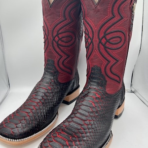 May include: A pair of cowboy boots with a striking design. The boots feature a combination of black and red tones, with a textured, snake-like pattern on the lower part and a smooth, burgundy upper. Intricate stitching in black and red adds detail.