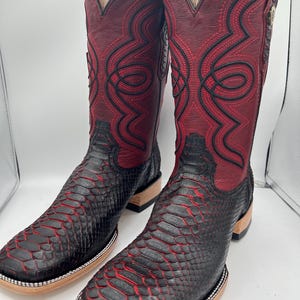 May include: A pair of cowboy boots with a striking design. The boots feature a combination of black and red colors, with a textured pattern on the lower part and intricate stitching on the upper shaft. The square-toe boots have a wooden sole.