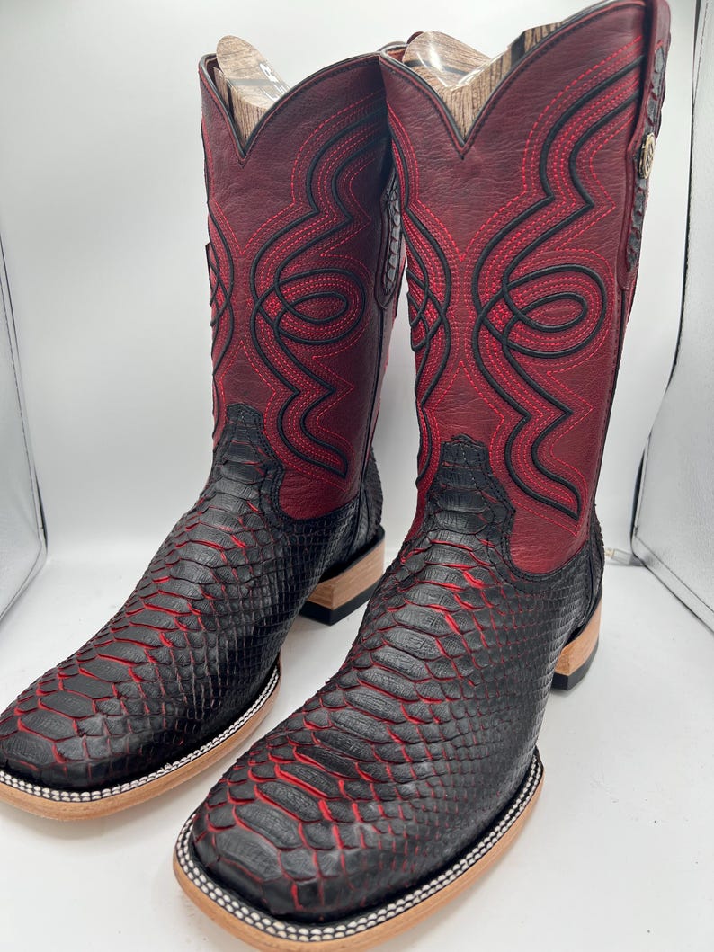 May include: A pair of cowboy boots with a striking design. The boots feature a combination of black and red colors, with a textured pattern on the lower part and intricate stitching on the upper shaft. The square-toe boots have a wooden sole.