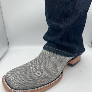 Peut inclure: Gros plan d'une botte de cowboy grise et blanche à motifs, avec un bout carré et une semelle marron clair. La botte est partiellement recouverte d'un jean bleu foncé.