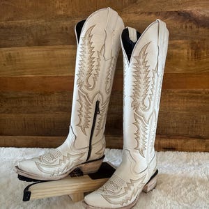 Könnte beinhalten: Ein Paar cremefarbene Cowboy-Stiefel mit aufwendigen Nähten und einer spitzen Zehenpartie. Die Stiefel haben einen hohen Schaft, einen seitlichen Reißverschluss und einen gestapelten Absatz. Die Stiefel werden auf einem Holzständer ausgestellt.