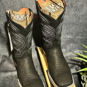 May include: A pair of black cowboy boots with a textured pattern and decorative stitching. The boots have a square toe and are displayed on a wooden stand. The interior of the boots is visible, with a light brown color.