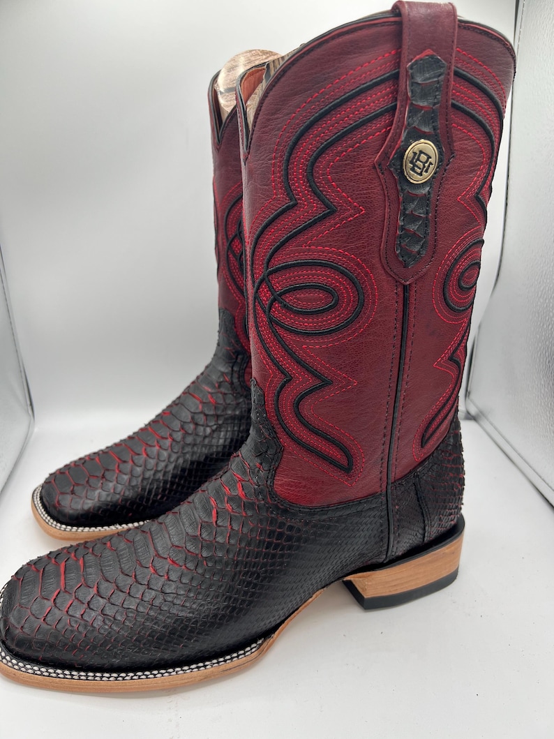 May include: A pair of cowboy boots with a striking design. The boots feature a combination of burgundy and black leather, with intricate red stitching and a snakeskin-like texture on the lower part. A gold emblem is visible on the top.