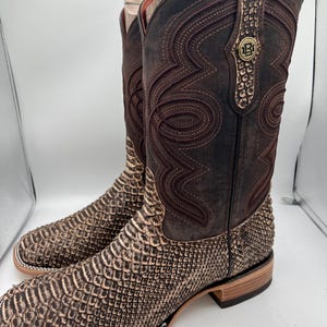 Puede incluir: Un par de botas de vaquero marrones con un estampado de piel de serpiente en el pie y la parte inferior de la pierna. La caña superior es de color marrón oscuro con costuras decorativas y un pequeño emblema metálico. Las botas tienen punta cuadrada y tacón apilado.