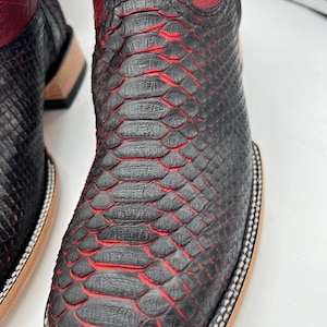May include: Close-up of a pair of black and red cowboy boots. The boots feature a textured, snake-like pattern with red accents. The top of the boot is red leather, and the sole has a decorative border.