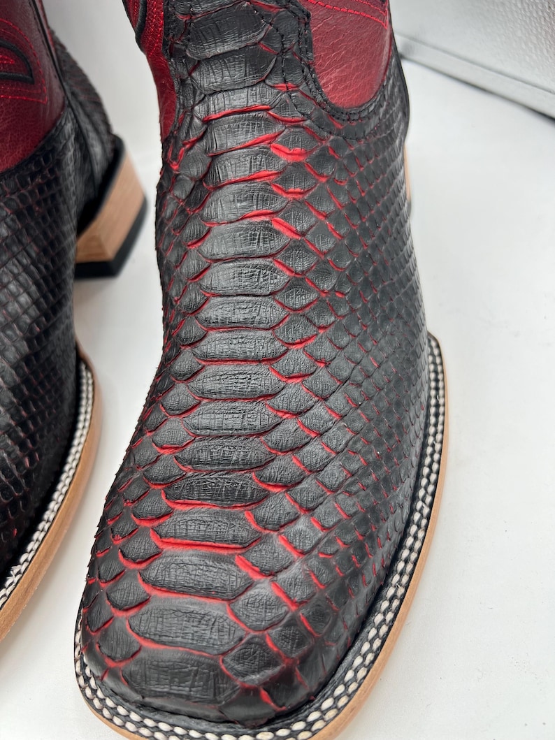 May include: Close-up of a pair of black and red cowboy boots. The boots feature a textured, snake-like pattern with red accents. The top of the boot is red leather, and the sole has a decorative border.
