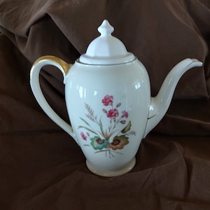 May include: White porcelain teapot with a gold rim and floral design. The teapot has a lid with a knob on top. The floral design features pink flowers, green leaves, and brown stems.