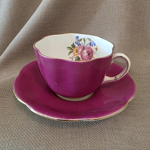 May include: A magenta teacup and saucer set with a scalloped edge and gold trim. The inside of the teacup features a floral design with pink and blue flowers. The set is placed on a neutral-colored surface.