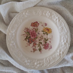 May include: White ceramic plate with a raised rim and a floral design. The design features pink, yellow, and brown flowers with green leaves.
